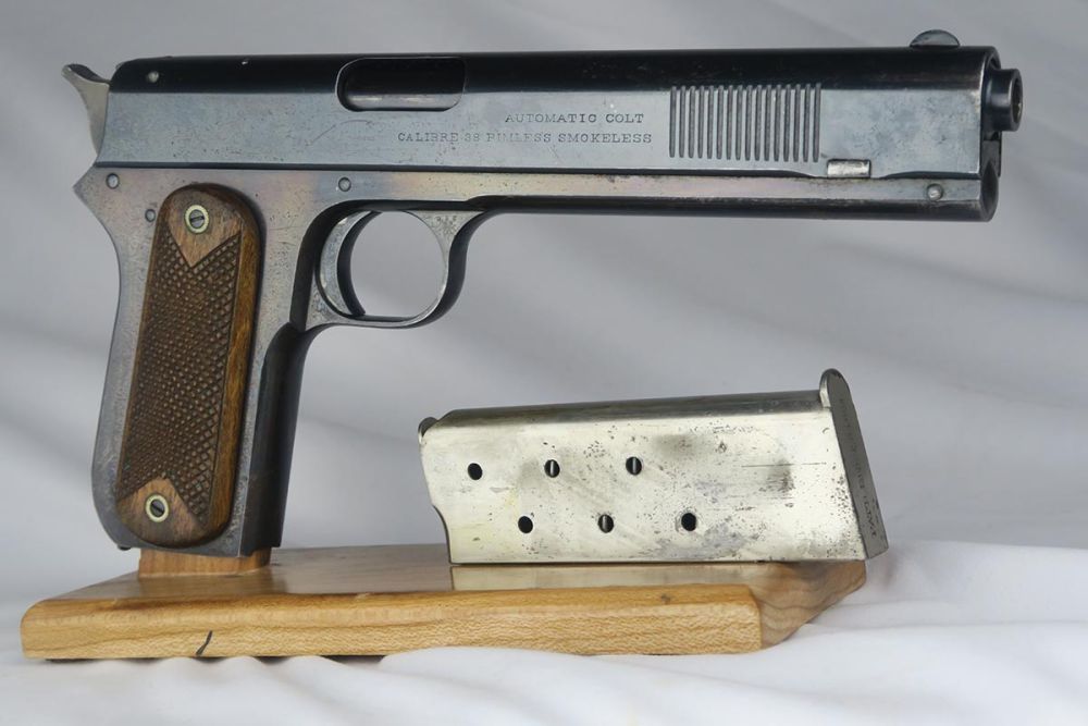 Colt Model 1900 semi-automatique en .38 ACP avec chargeur, présenté sur un socle en bois.