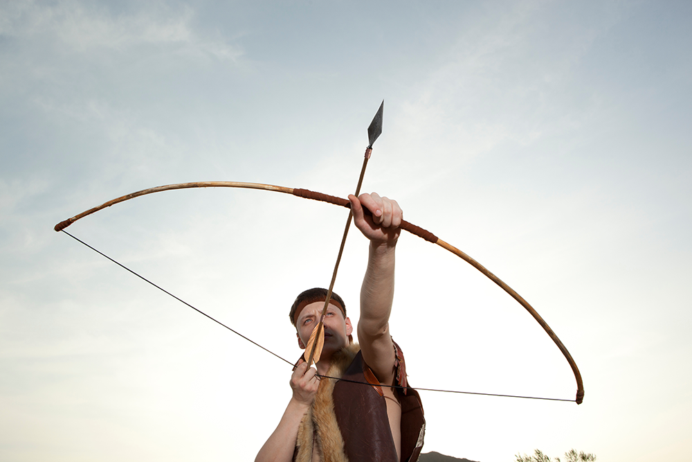 Un archer en tenue primitive tire une flèche avec un arc long en bois, face au ciel.