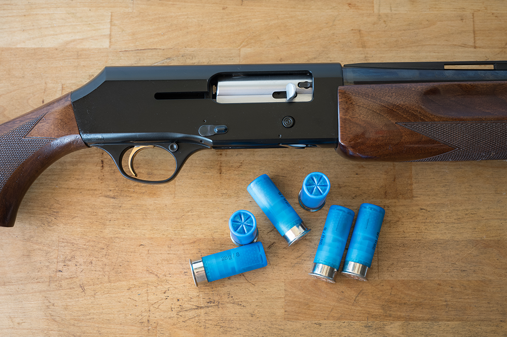 Fusil de chasse semi-automatique calibre 12 avec culasse ouverte, posé sur une table en bois aux côtés de cartouches bleues