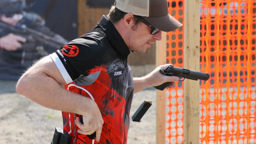 Tireur IPSC rechargeant un pistolet 1911 en match, devant une barrière orange de sécurité.