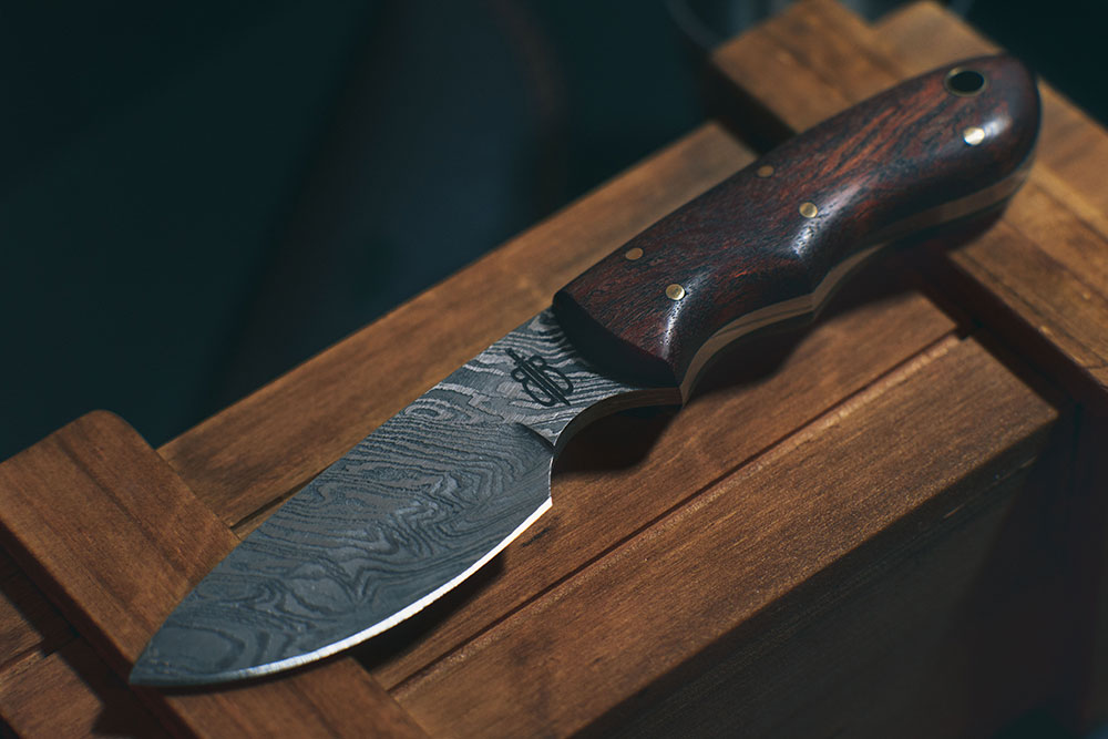Couteau fixe à lame Drop point en acier Damas, manche en bois exotique, posé sur une boîte en bois.