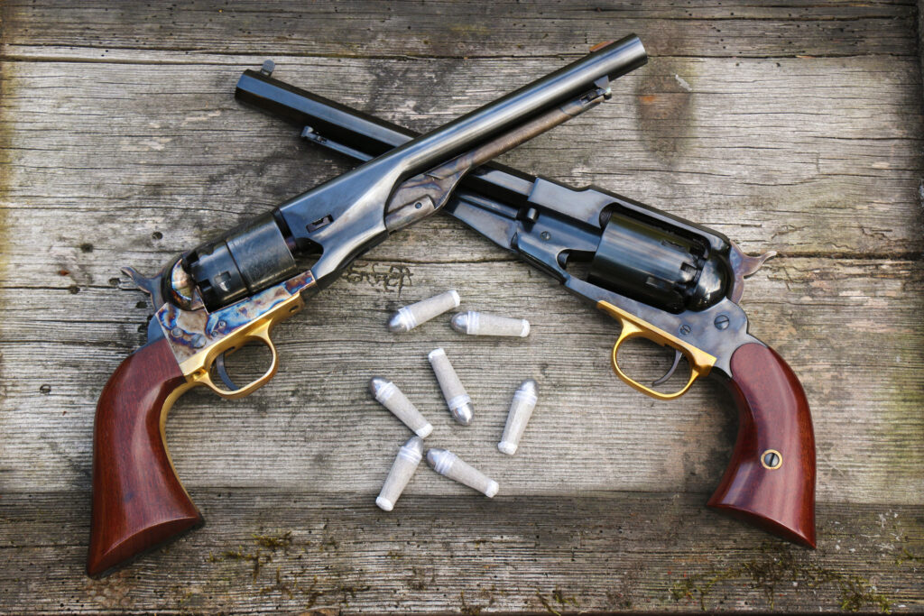 Deux revolvers Remington1858 à poudre noire croisés sur une table en bois, avec balles en plomb calibre .44 disposées au centre.