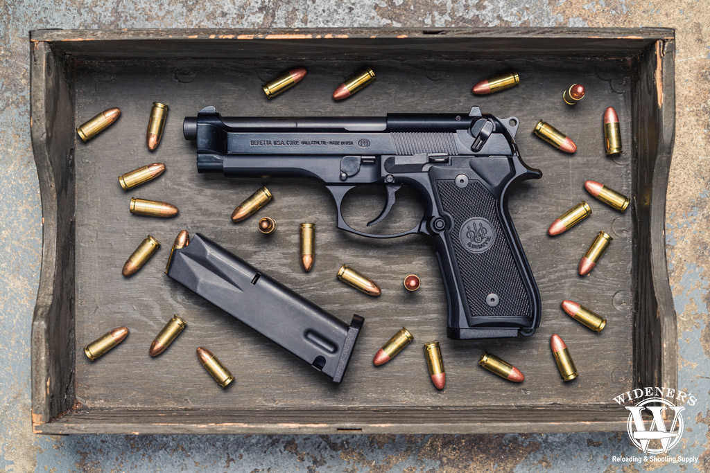 Photo d’un pistolet Beretta 92 noir avec son chargeur détaché et des cartouches 9x19 mm dispersées autour, posé dans un coffret en bois.

