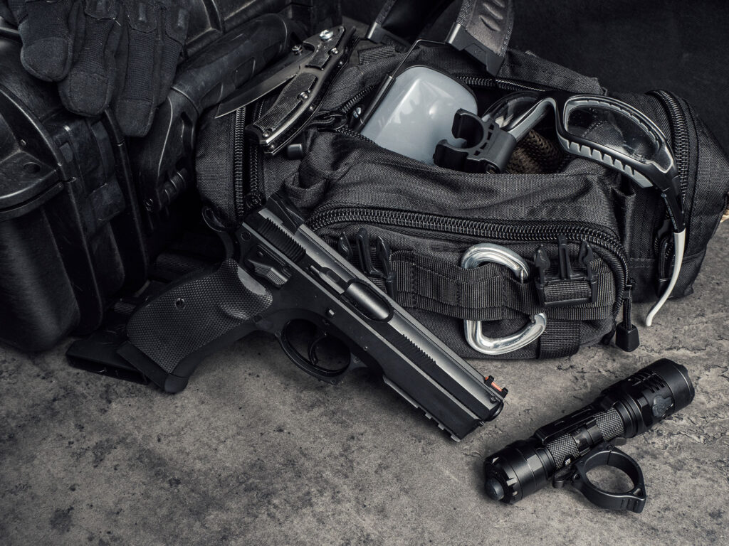 Tireur préparant son sac de tir avec pistolet, lampe, lunettes de protection et accessoires organisés sur le sol.