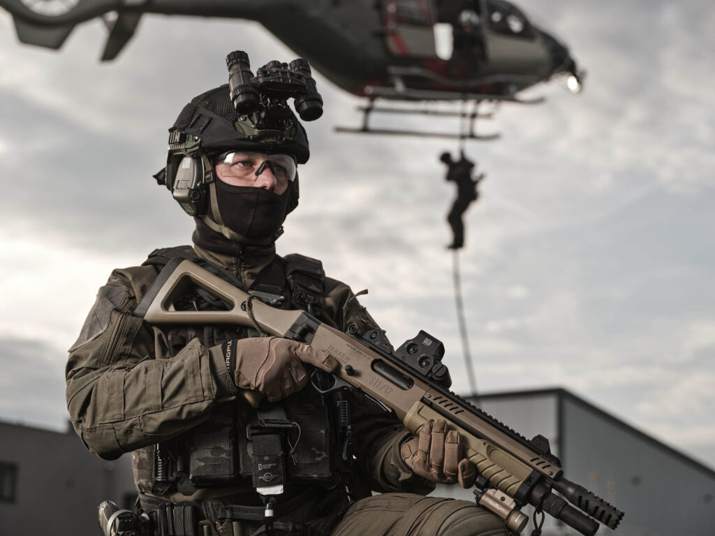 Opérateur équipé d’un fusil Fabarm STF12 tactique, portant casque avec vision nocturne et gilet tactique, avec un hélicoptère en arrière-plan lors d’une intervention.