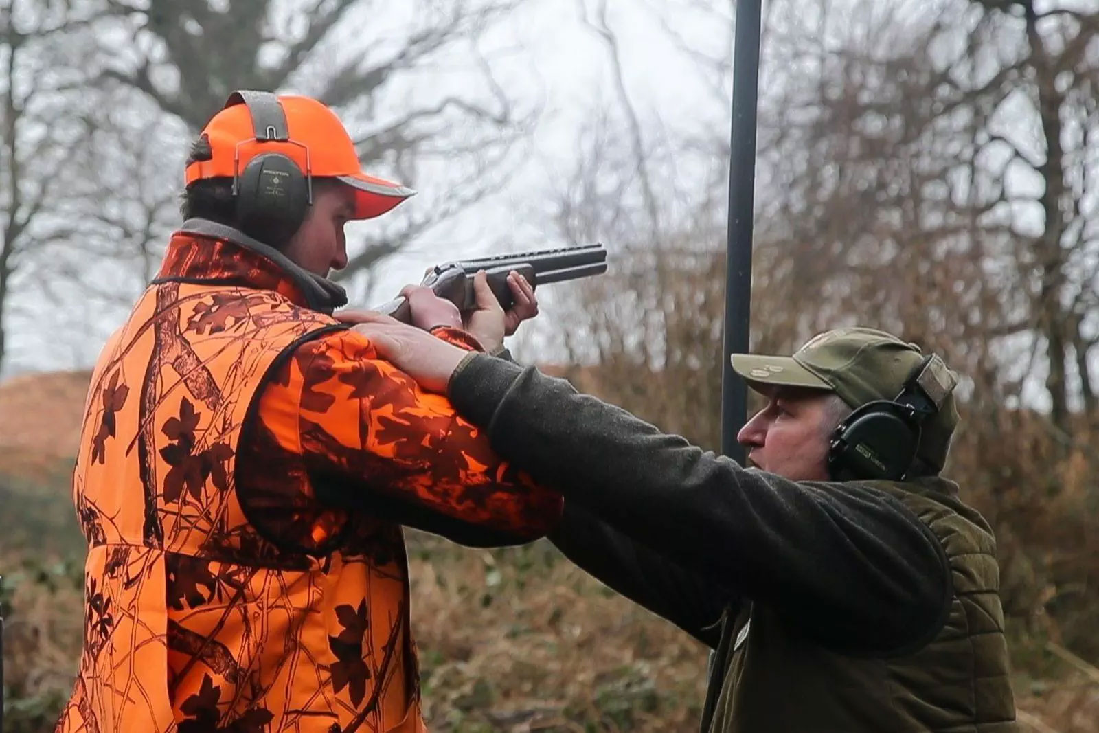 Un instructeur guide un candidat portant un gilet de sécurité orange lors de l'examen pratique du permis de chasser, avec un fusil pointé en position de tir.