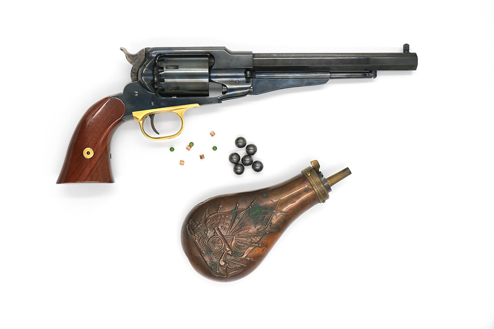 Revolver Remington 1858 New Army à poudre noire avec poire à poudre, balles en plomb et amorces de percussion sur fond blanc.