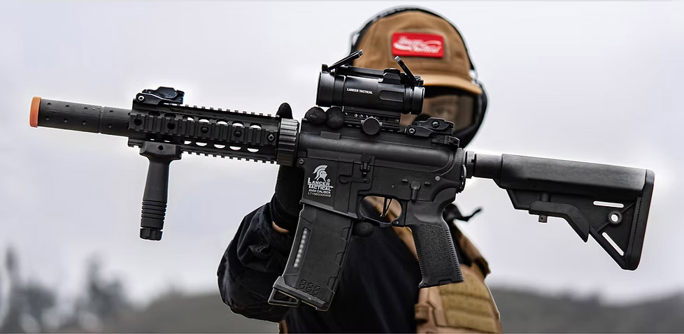 Joueur d’airsoft utilisant une réplique Lancer Tactical Gen 3 équipée d’un viseur point rouge et d’accessoires tactiques lors d’une partie en extérieur.