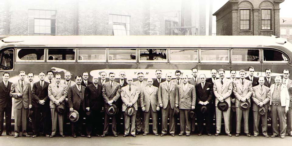 Photo historique d’un groupe d’hommes devant un autocar et les bâtiments de l’entreprise Mossberg